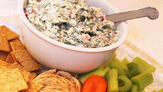 A bowl of spinach dip next to some crackers and vegetables.